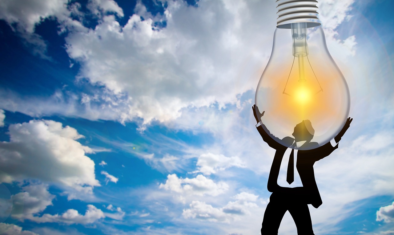 A silhouetted figure raising a large glowing lightbulb above their head against a blue sky.